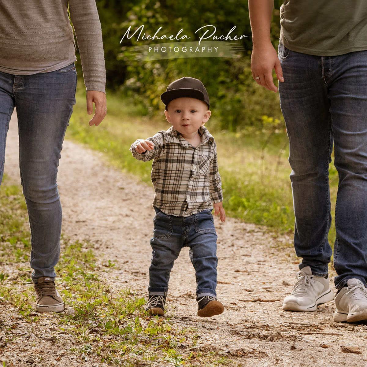 Kleiner Junge mit Kappe geht selbstbewusst zwischen seinen Eltern einen sonnigen Waldweg entlang.